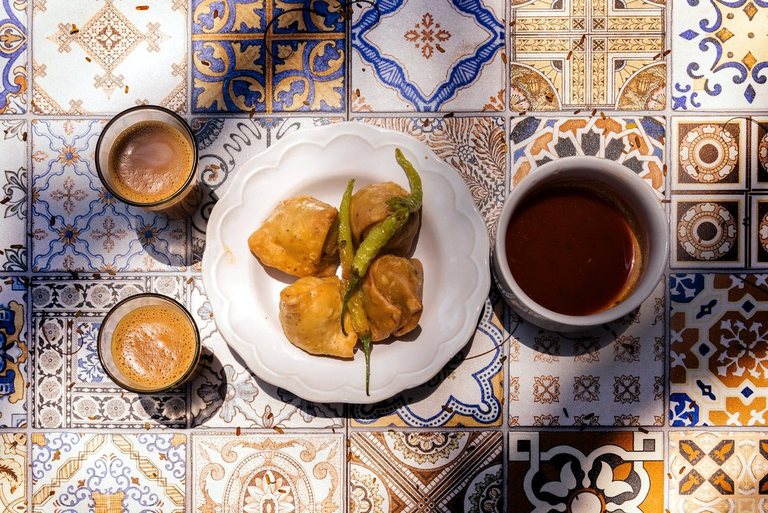 Samosas and a fried green chilli on a plate, with glasses of chai, on patterned tiles
